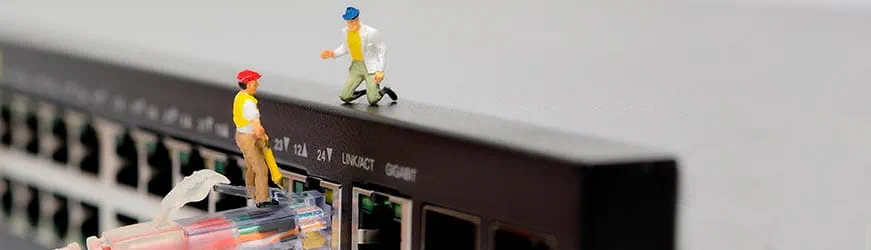 Deux figurines représentant des techniciens en télécommunication, l’un, portant un casque rouge, debout sur une prise RJ45, l’autre, avec un casque bleu, à genoux, sur une box au-dessus des prises.