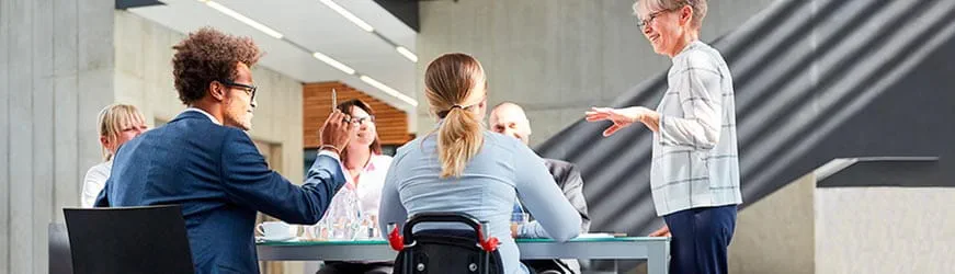 Six personnes dans un bureau, autour d’une table pendant une réunion. Une des personnes prenant part à la discussion est sur un fauteuil roulant, de dos