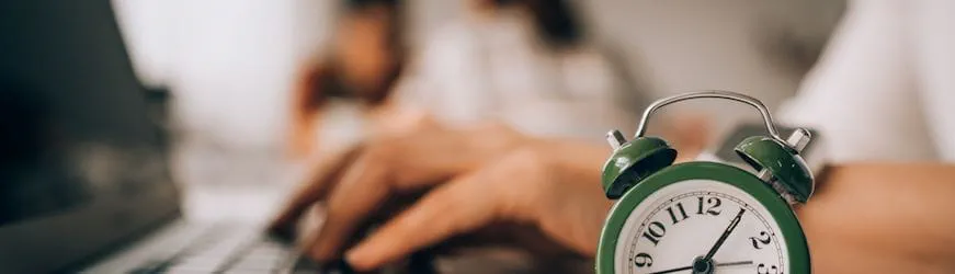 Les mains d’un salarié qui tapent sur son clavier d’ordinateur, à côté d’un réveil qui indique que ce sera bientôt le moment de sa pause