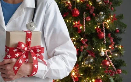 Un soignant tenant un cadeau devant un sapin de Noël illuminé, illustrant un Secret-Santa dans le secteur de la santé.