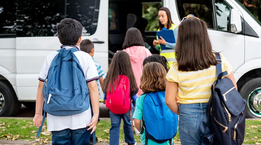 Niños y niñas de espalda con mochilas acercándose al autobús escolar