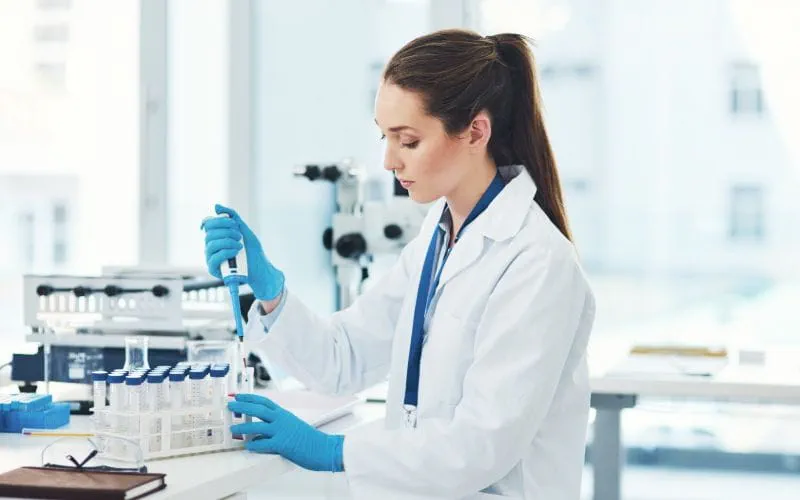Technicienne de laboratoire en blouse blanche manipulant une pipette pour effectuer des analyses dans un environnement scientifique, entourée de tubes à essai.