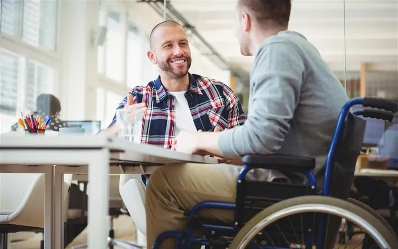 Un homme souriant et barbu, vêtu d'une chemise à carreaux, discute avec une personne en fauteuil roulant Un homme souriant et barbu, vêtu d'une chemise à carreaux, discute avec une personne en fauteuil roulant