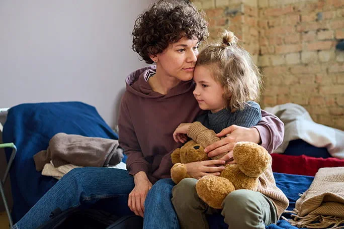 Une femme assise réconforte tendrement une petite fille tenant un ours en peluche, dans un environnement simple et chaleureux, illustrant un moment de soutien et de lien affectif.