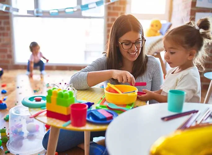 Une enseignante souriante avec des lunettes aide une petite fille à jouer avec des jouets colorés à une petite table