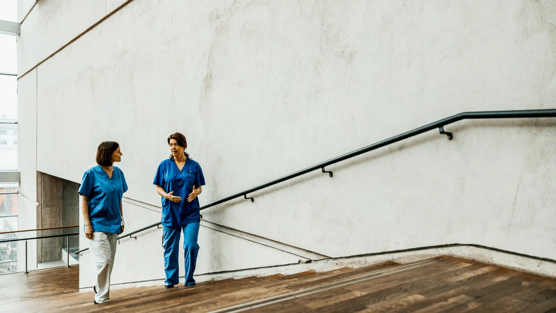 Two healthcare professionals in blue scrubs walk together and converse on a wooden staircase in a bright, modern hospital corridor.
