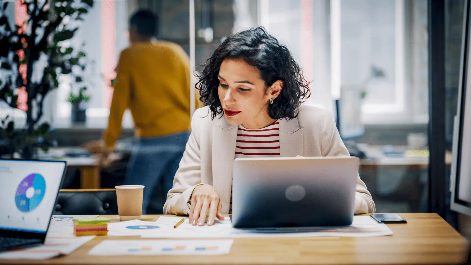 Une femme aux cheveux courts et foncés est assise à son bureau près d’un ordinateur portable ouvert, examinant des graphiques imprimés disposés sur la table. Une femme aux cheveux courts et foncés est assise à son bureau près d’un ordinateur portable ouvert, examinant des graphiques imprimés disposés sur la table.