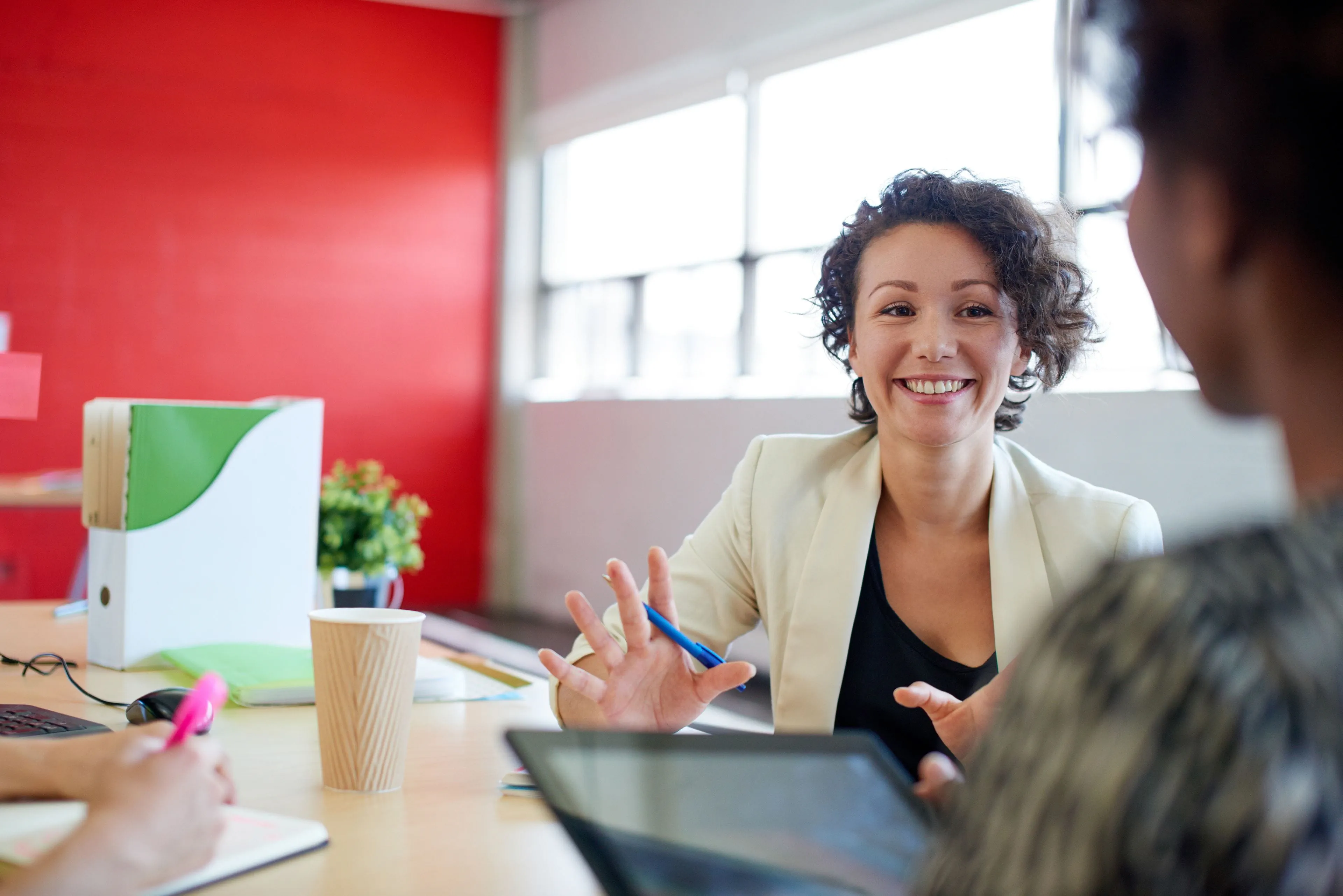 Femme assise à une table de salle de réunion dans un environnement de travail, en train de parler avec une collègue.