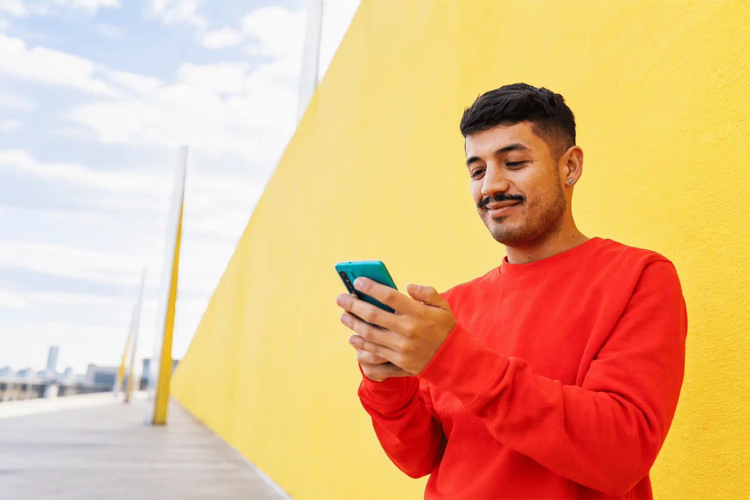 Person wearing a red sweater standing in front of a yellow wall while looking at a smartphone. Architectural background on the left provides ample negative space for website content.