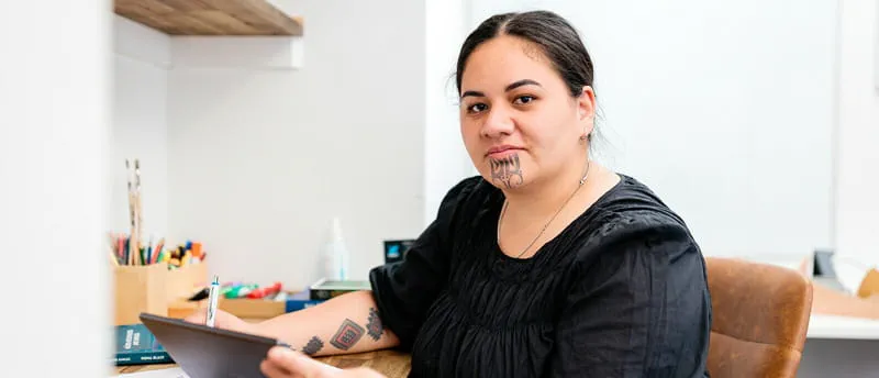 Person with Māori tāmoko tattoos working at a desk with a pen and tablet in an office setting