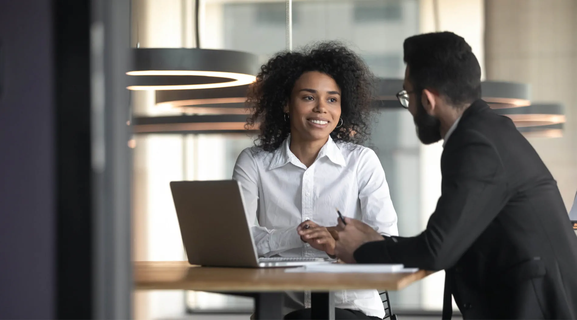 Two people in professional attire having a discussion at a table with a laptop in an office setting