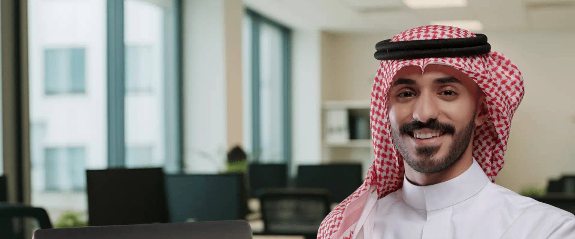 Smiling Saudi man in traditional attire working on a laptop in a modern office environment