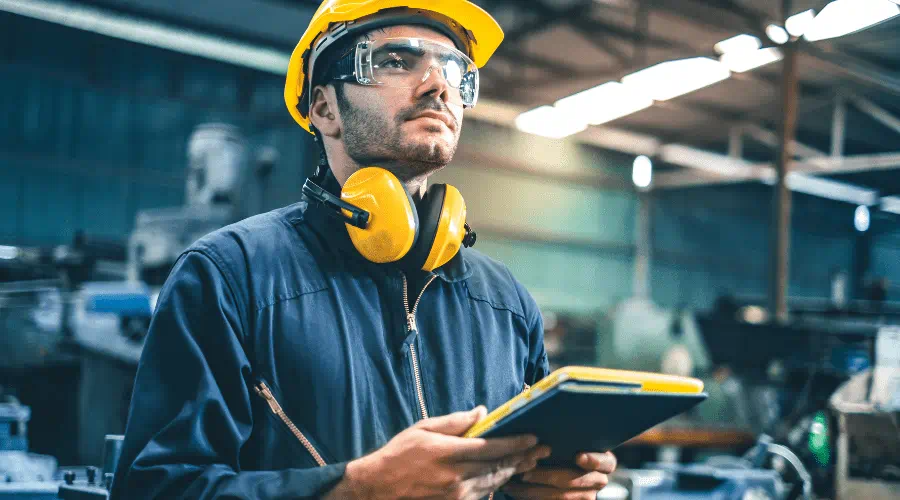 Industriarbetare i skyddsutrustning,  håller en surfplatta och inspekterar  produktionsmiljön