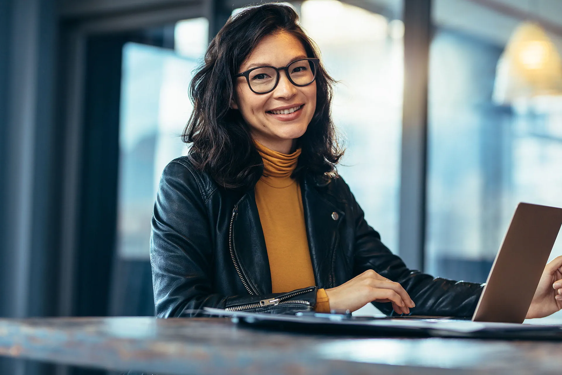 Smiling woman with glasses and a black leather jacket working on a laptop at a wooden table in a modern, well-lit office or cafe with large windows.