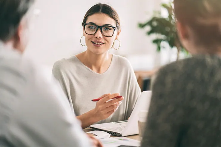 Smiling woman with glasses holding a pen and engaging in a discussion with two people in an office setting, focusing on collaboration and teamwork. Smiling woman with glasses holding a pen and engaging in a discussion with two people in an office setting, focusing on collaboration and teamwork.