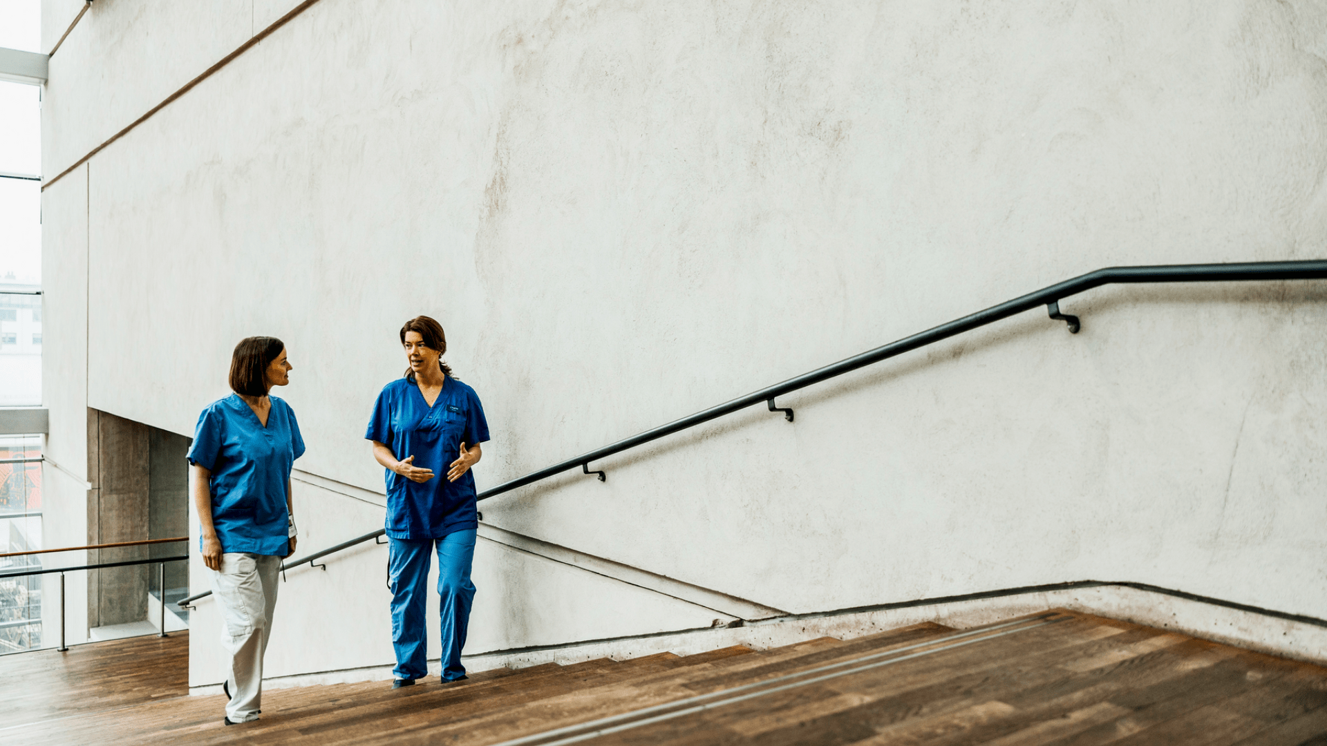 Two healthcare professionals in blue scrubs walk together and converse on a wooden staircase in a bright, modern hospital corridor.
