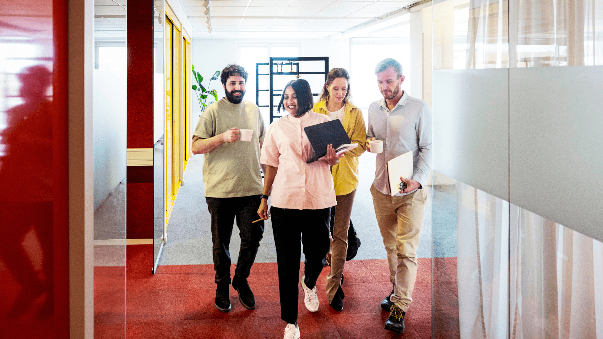A group of five coworkers walking together through a bright office hallway, smiling and talking