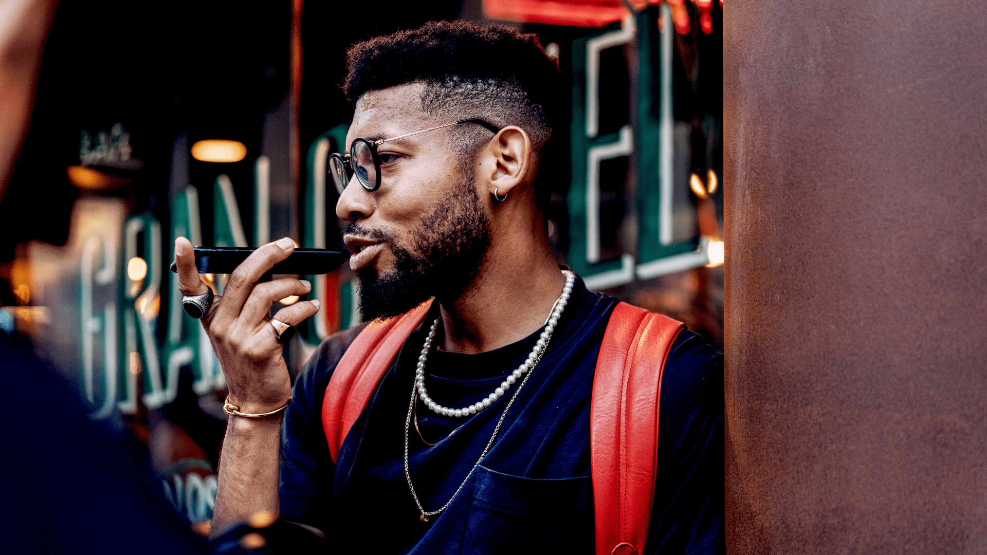 man wearing glasses and a red backpack speaking on the phone outside