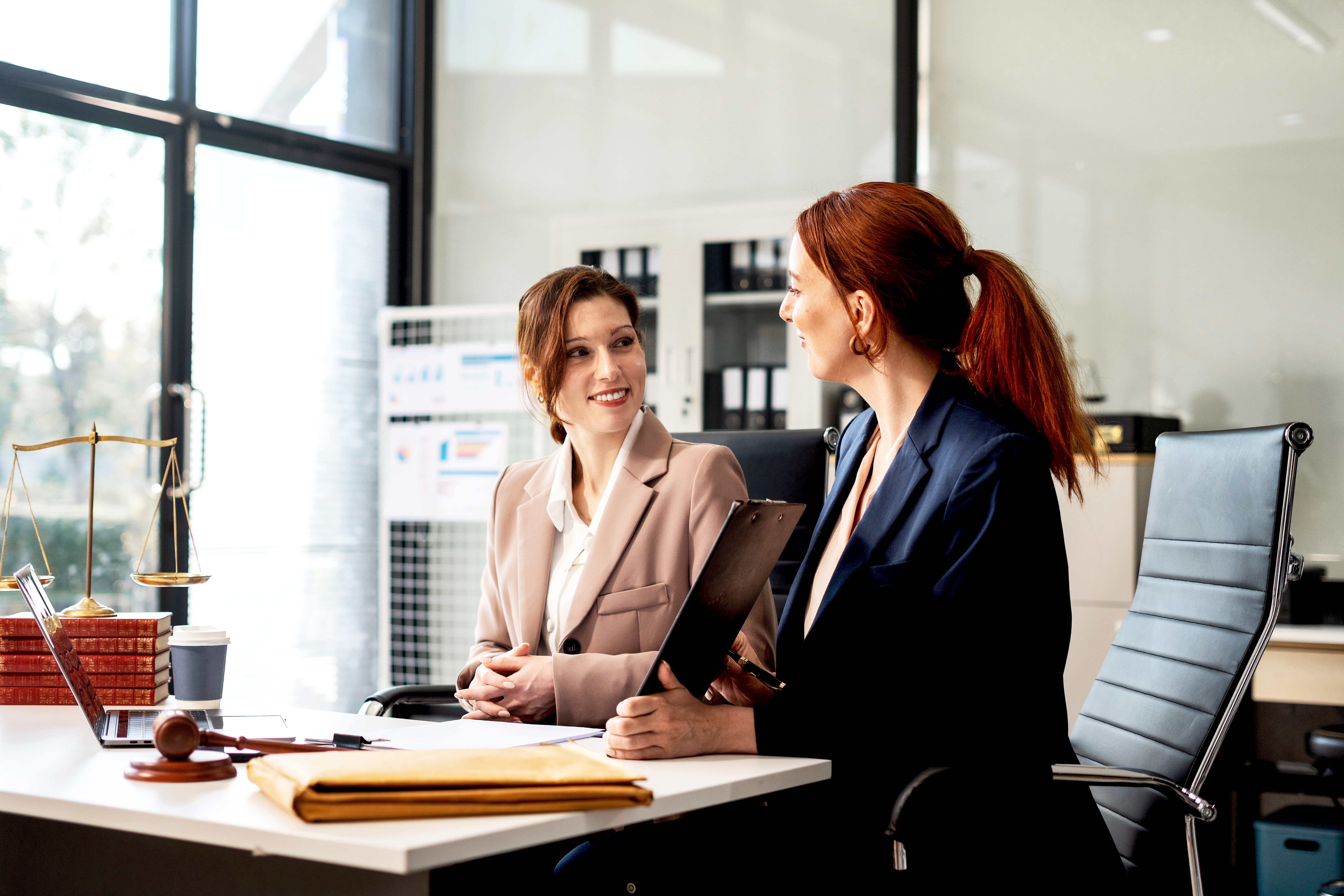 two women dressed in suits sitting at a desk in a work office, smiling