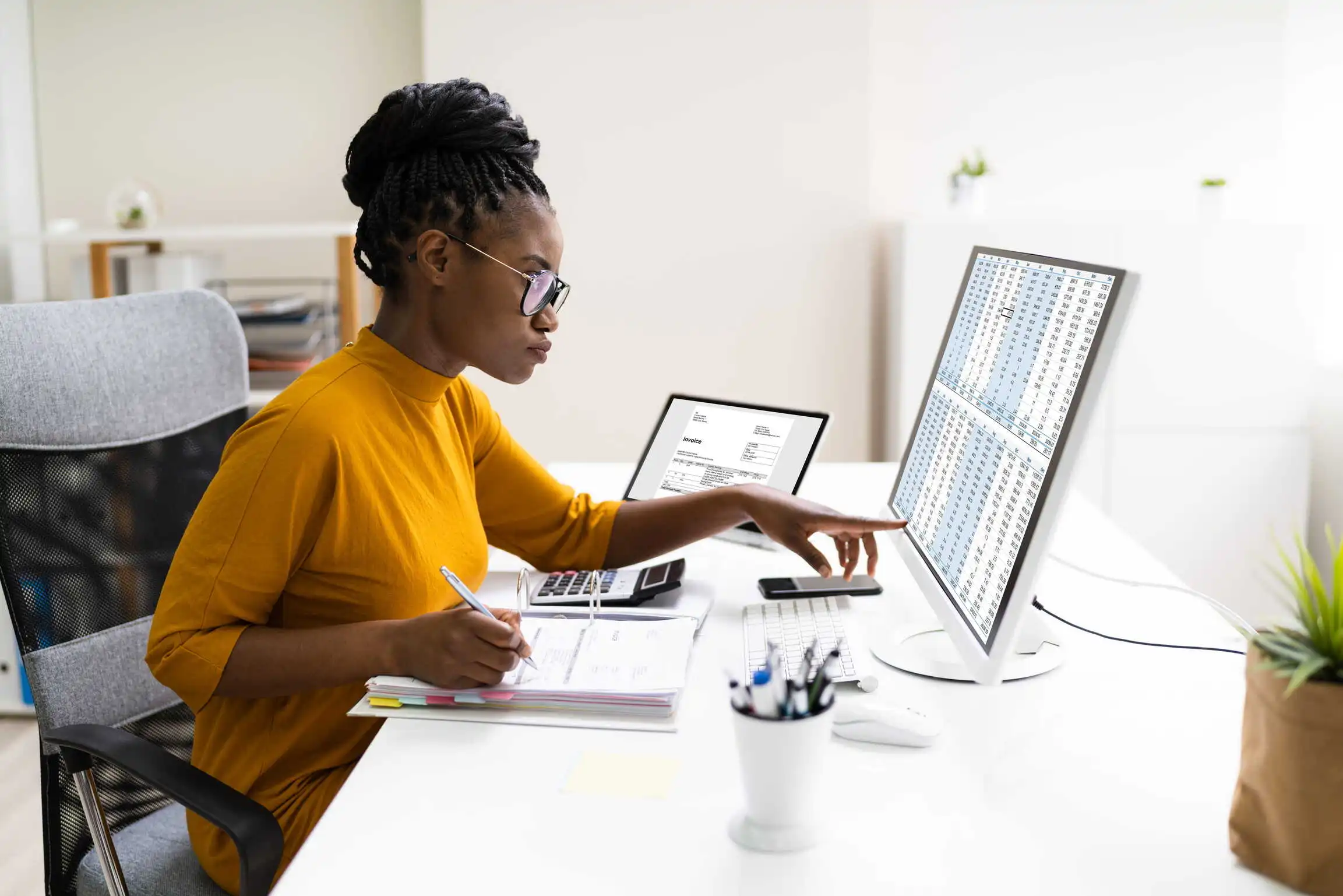 Female manager scrutinizes her department’s budget and personnel costs across multiple screens, documents, and calculators on her desk.