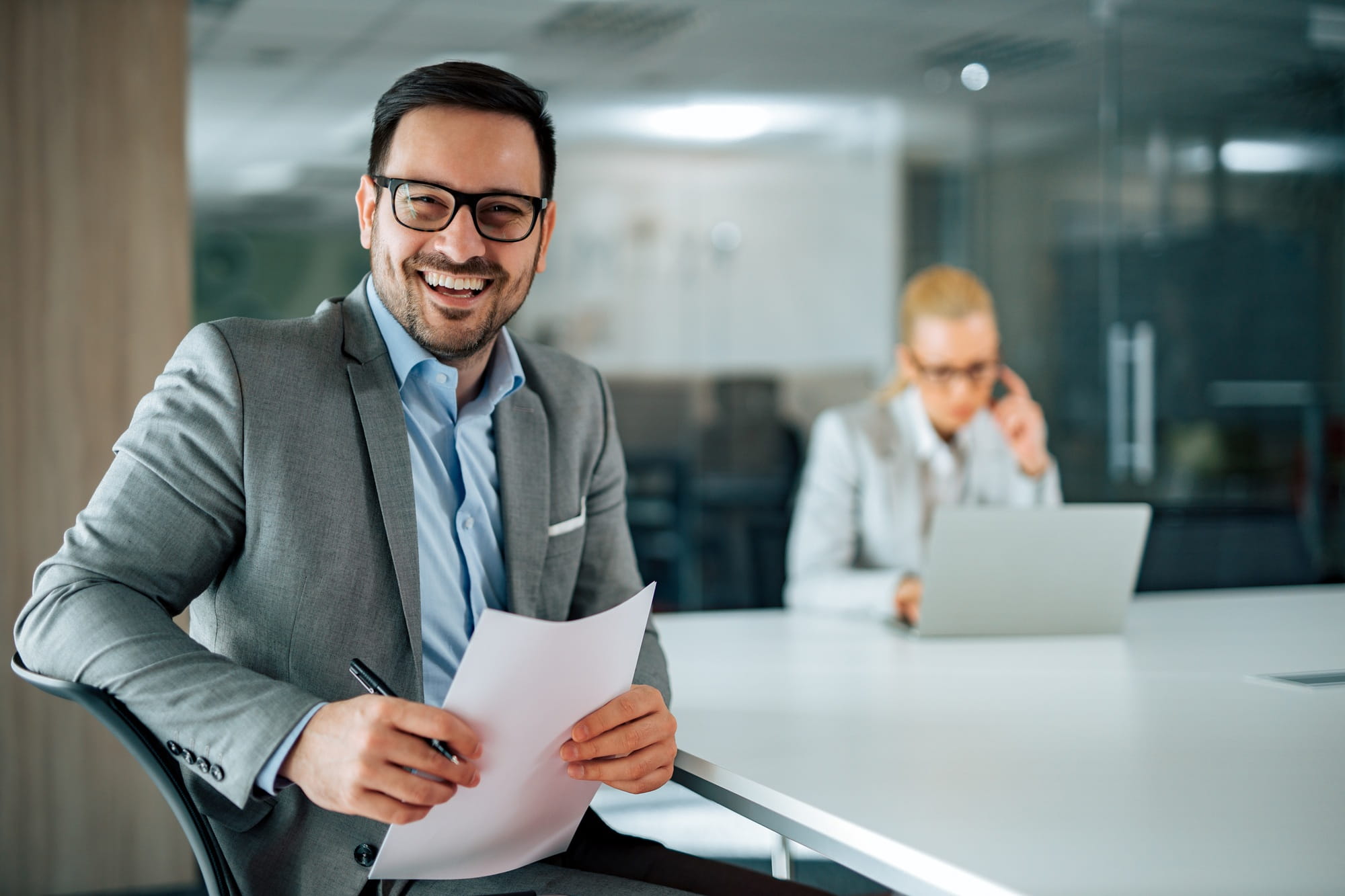 Imprenditore sorridente con un foglio in mano, collega femminile sullo sfondo Imprenditore sorridente con un foglio in mano, collega femminile sullo sfondo