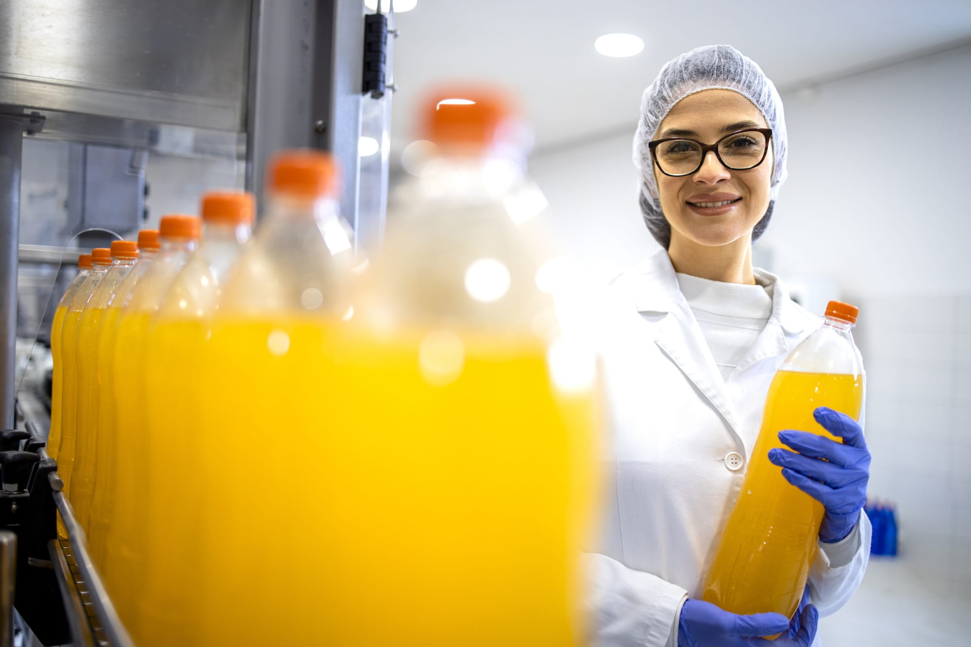Una donna con camice bianco e cuffia sterile tiene in mano una bottiglia con una bevanda gialla in uno stabilimento di produzione. Una donna con camice bianco e cuffia sterile tiene in mano una bottiglia con una bevanda gialla in uno stabilimento di produzione.