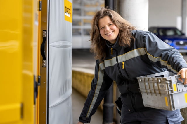 A postal worker in uniform loads a plastic crate into a yellow delivery vehicle at a logistics station. A postal worker in uniform loads a plastic crate into a yellow delivery vehicle at a logistics station.