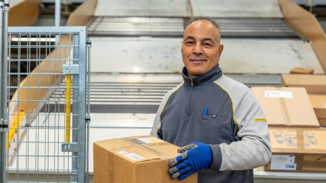 Person carrying a package in a parcel sorting facility, standing near a conveyor system and surrounded by boxes and metal structures. Person carrying a package in a parcel sorting facility, standing near a conveyor system and surrounded by boxes and metal structures.