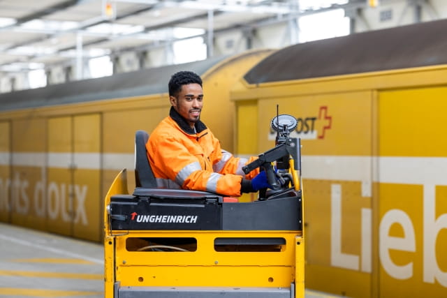 Person driving a yellow industrial vehicle in a logistics facility, positioned in front of a yellow postal transport container. Person driving a yellow industrial vehicle in a logistics facility, positioned in front of a yellow postal transport container.