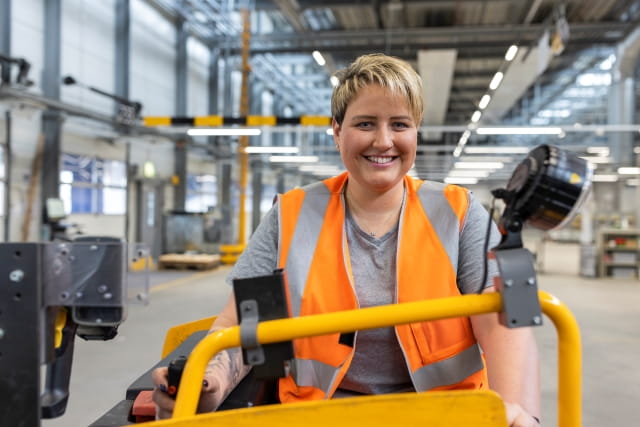 Person wearing an orange safety vest operating an industrial vehicle inside a large warehouse or logistics facility. Person wearing an orange safety vest operating an industrial vehicle inside a large warehouse or logistics facility.