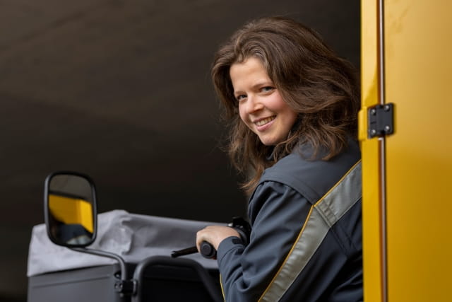 Person in work attire operating a delivery vehicle, looking to the side next to the vehicle’s yellow door. Person in work attire operating a delivery vehicle, looking to the side next to the vehicle’s yellow door.
