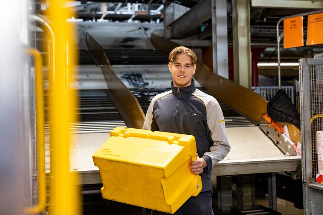 An employee holds a yellow transport box in front of an automated sorting system inside a logistics facility. An employee holds a yellow transport box in front of an automated sorting system inside a logistics facility.