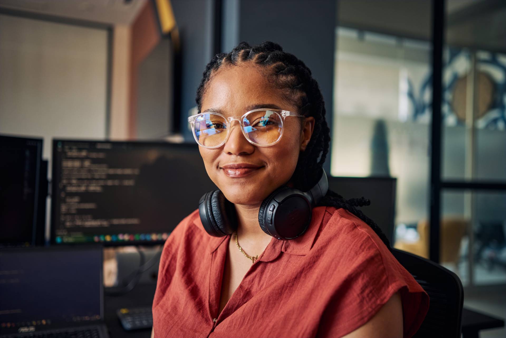 Foto einer lächelnden Frau mit geflochtenen Haaren und Brille an einem Arbeitsplatz. Sie trägt ein rotes Oberteil und Kopfhörer um den Hals; im Hintergrund sind Monitore mit unscharfem Code in einem modernen Büro zu sehen.