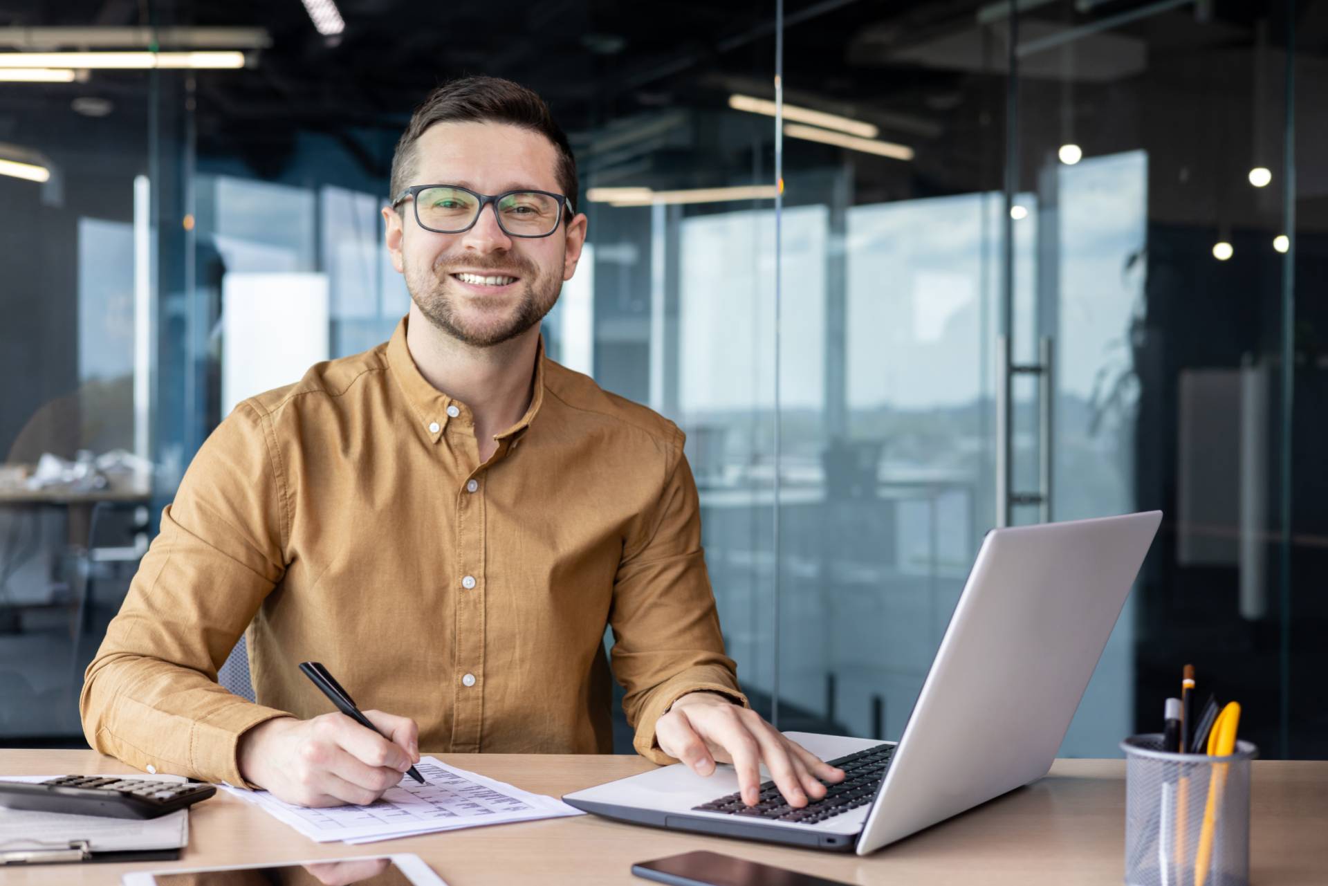 Mann sitzt in einem modernen Büro am Schreibtisch und arbeitet am Laptop. Er lächelt in die Kamera. Auf dem Tisch liegen Rechner, Dokumente und Stifte.