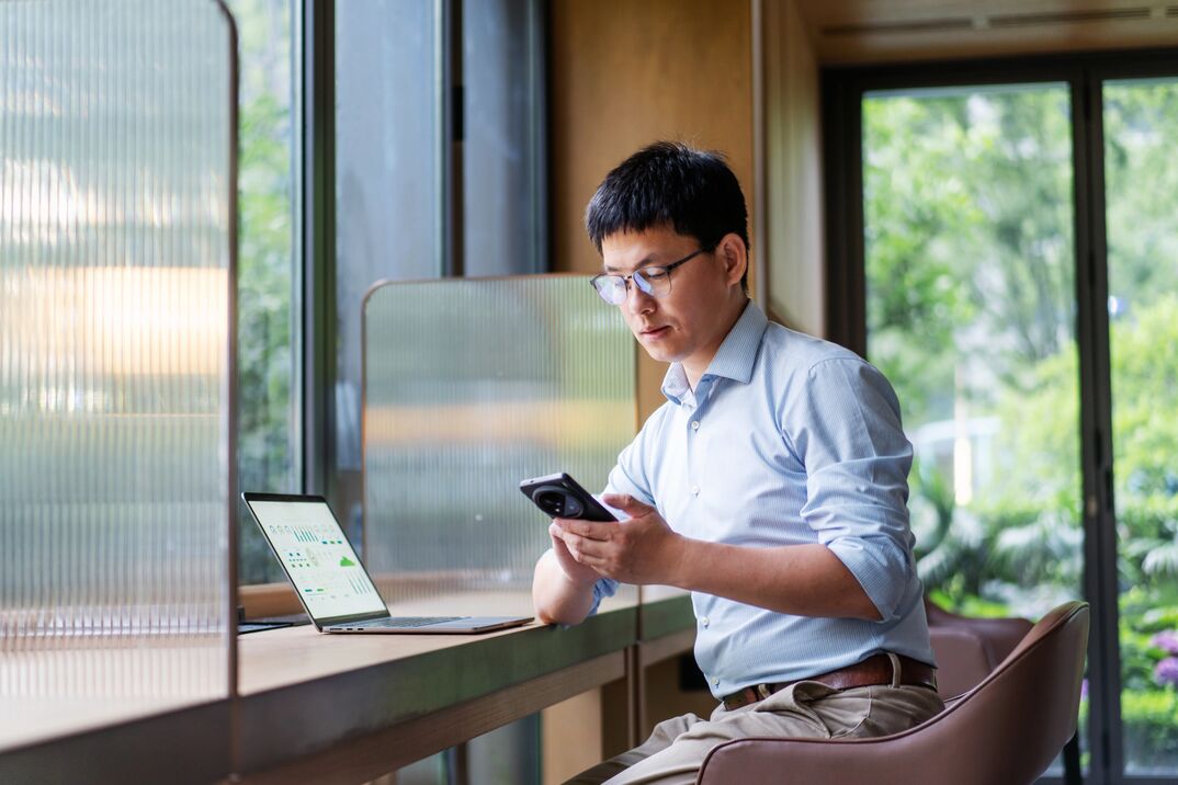Person using a smartphone while working at a laptop in a modern office.