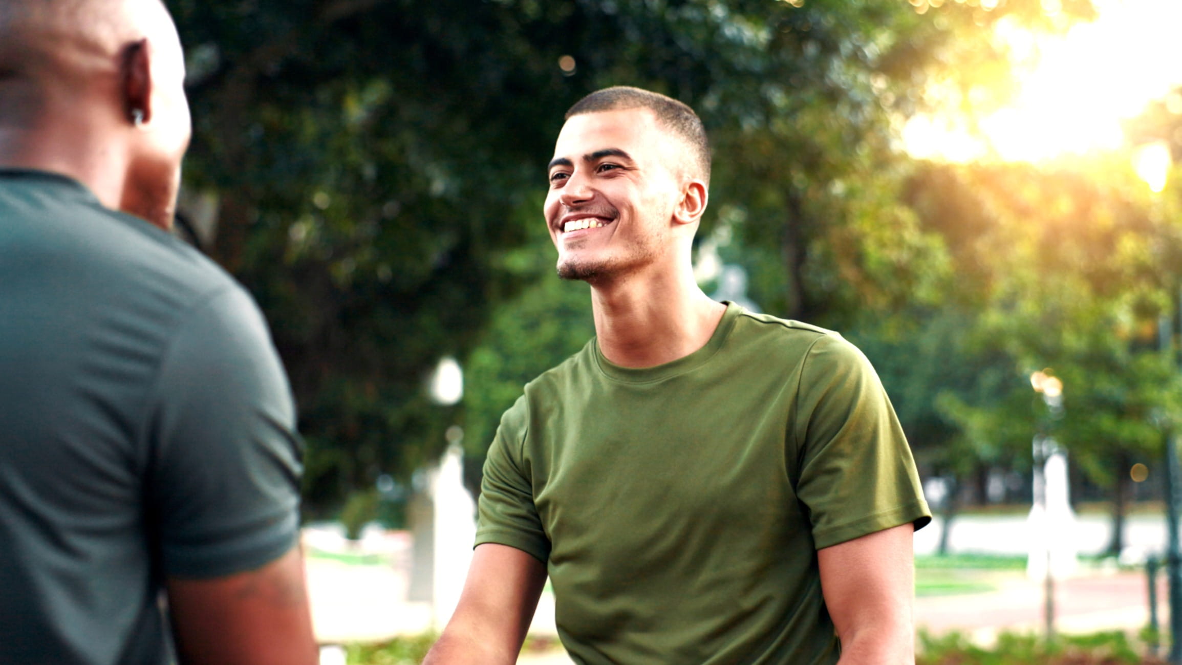 Two people talking outdoors in a park with trees and sunlight in the background.