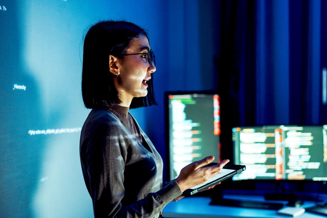 Person holding a tablet in a dim office with multiple monitors displaying code.