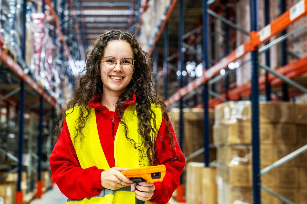 Person wearing a high-visibility safety vest and red jacket holding a handheld scanner while standing in a warehouse