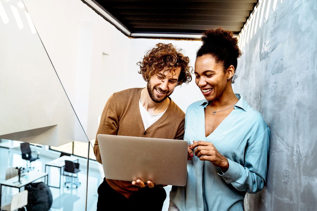 Two professionals standing in a modern office hallway reviewing information together on a laptop.