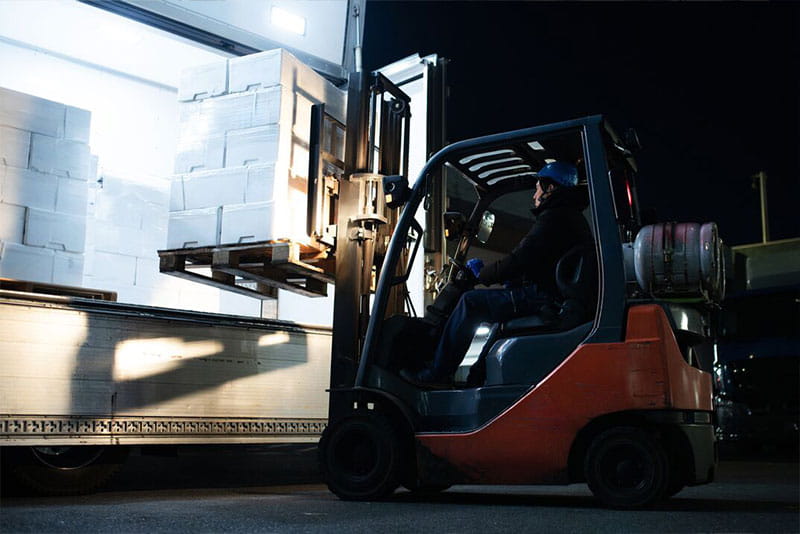 Forklift operator loading pallets onto a delivery truck at a busy warehouse during peak season.