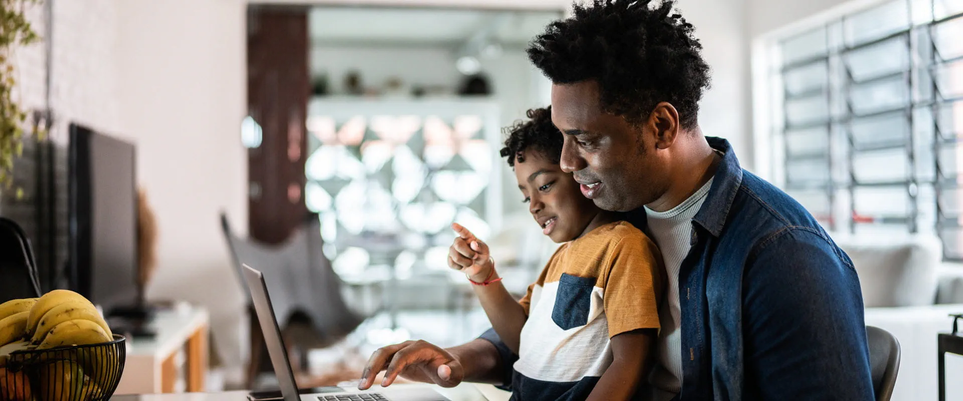 Father and young son sitting together at a desk, smiling as they look at a laptop screen in a cozy home setting.