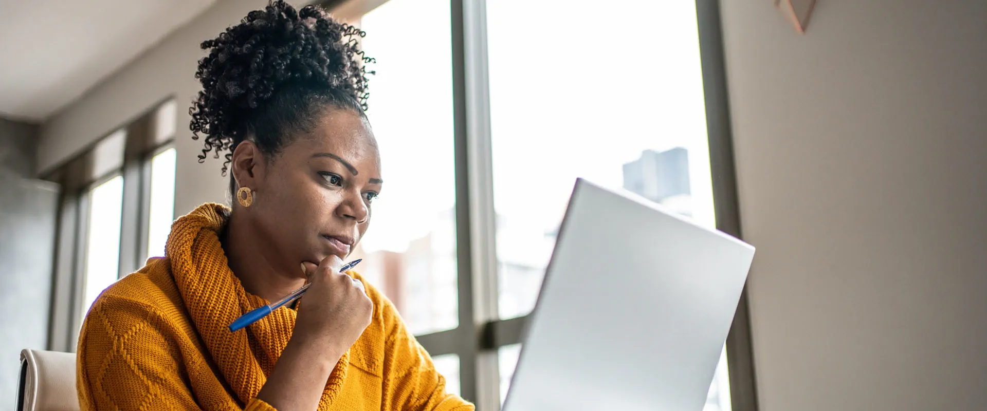 Focused professional working on a laptop at home, wearing a yellow sweater, holding a pen
