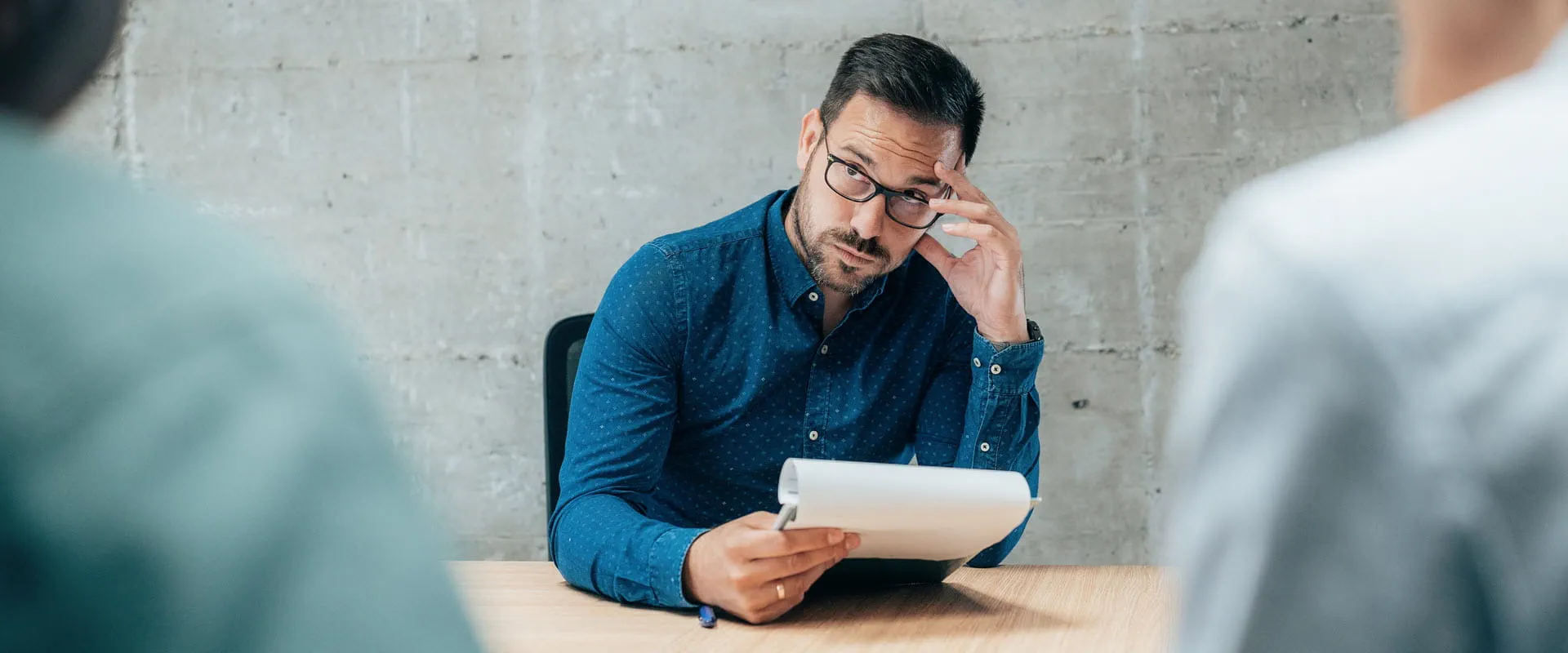 Person in a job interview holding a notepad, appearing thoughtful while listening to two interviewers.
