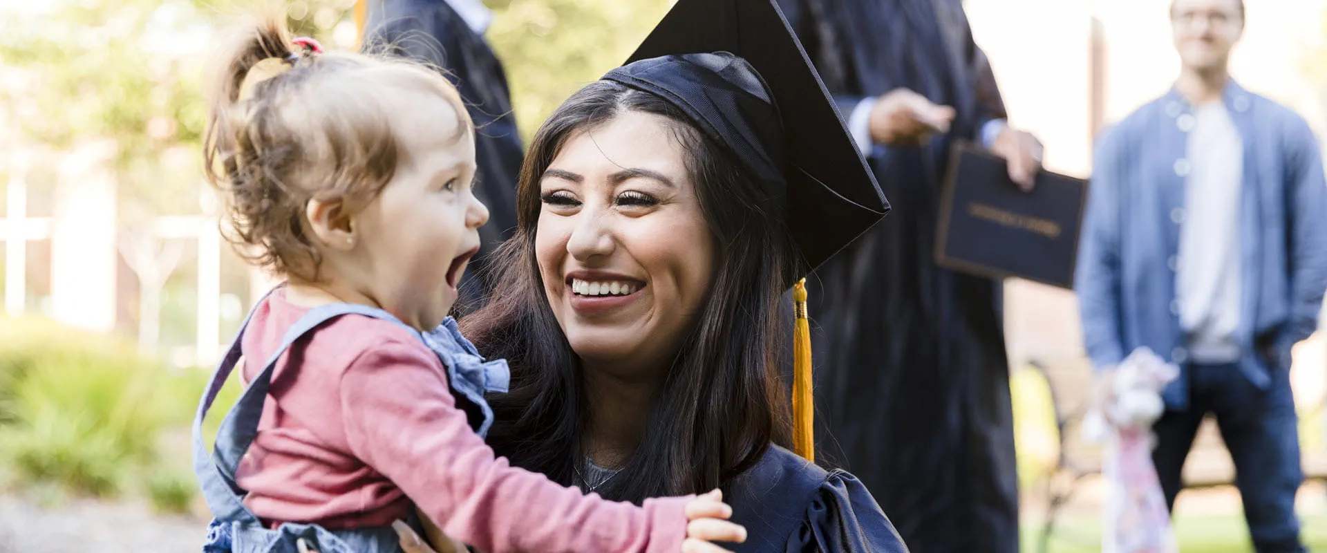 A parent in graduation robe smiles while holding their young toddler.