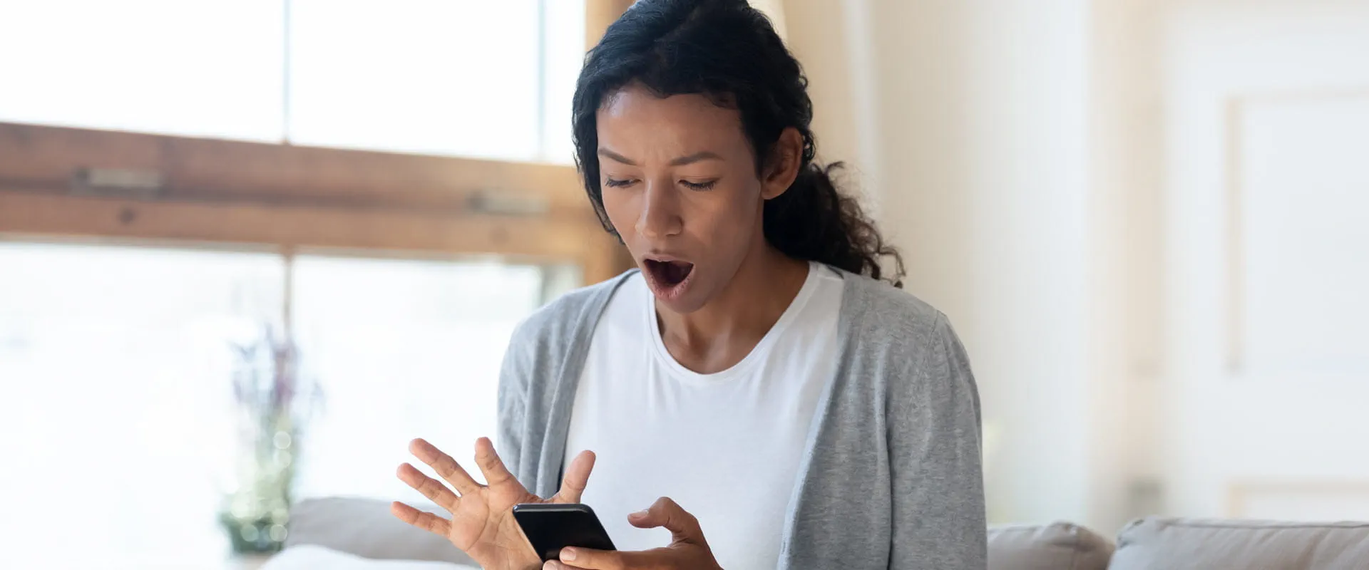 A young person in a t-shirt and cardigan sits on a couch at home. They look shocked as they stare at the mobile phone in their hand.