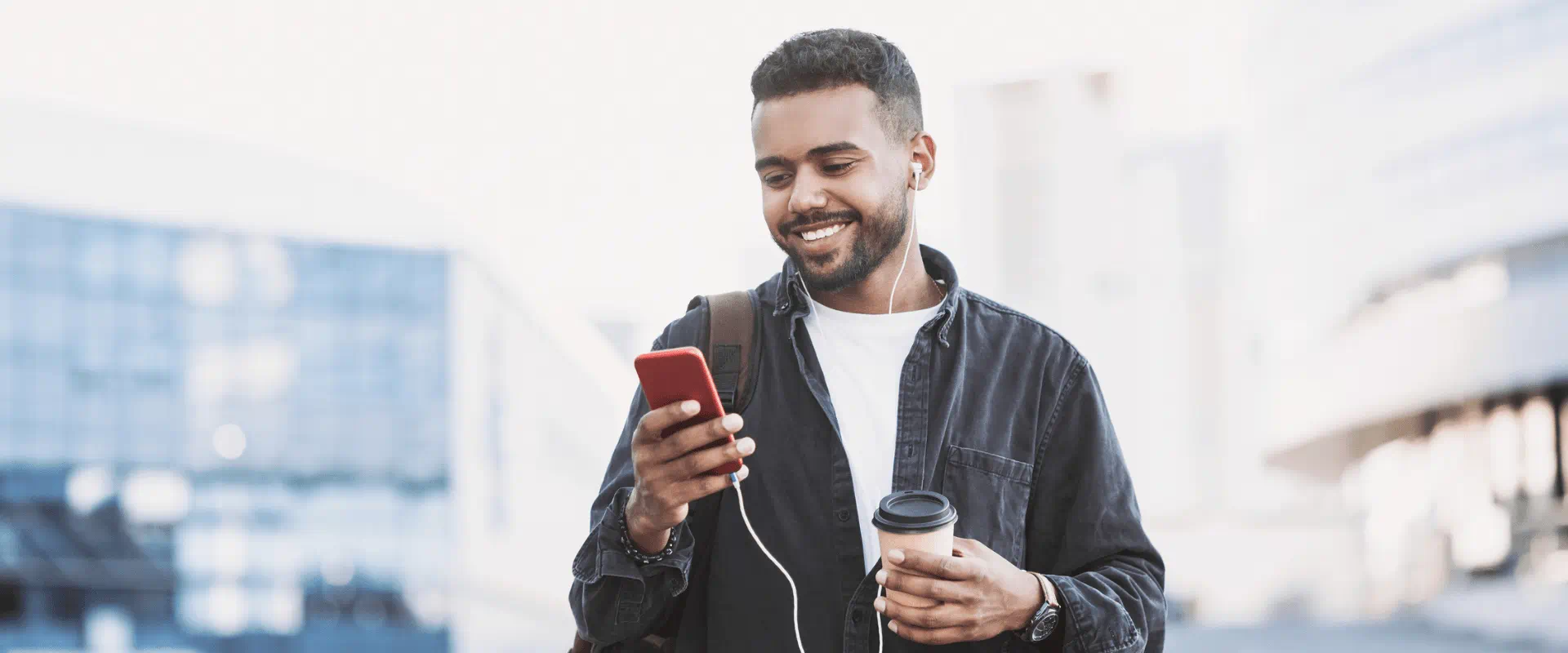 A person outside, wearing earbuds, looks at their phone while holding a cup of coffee.