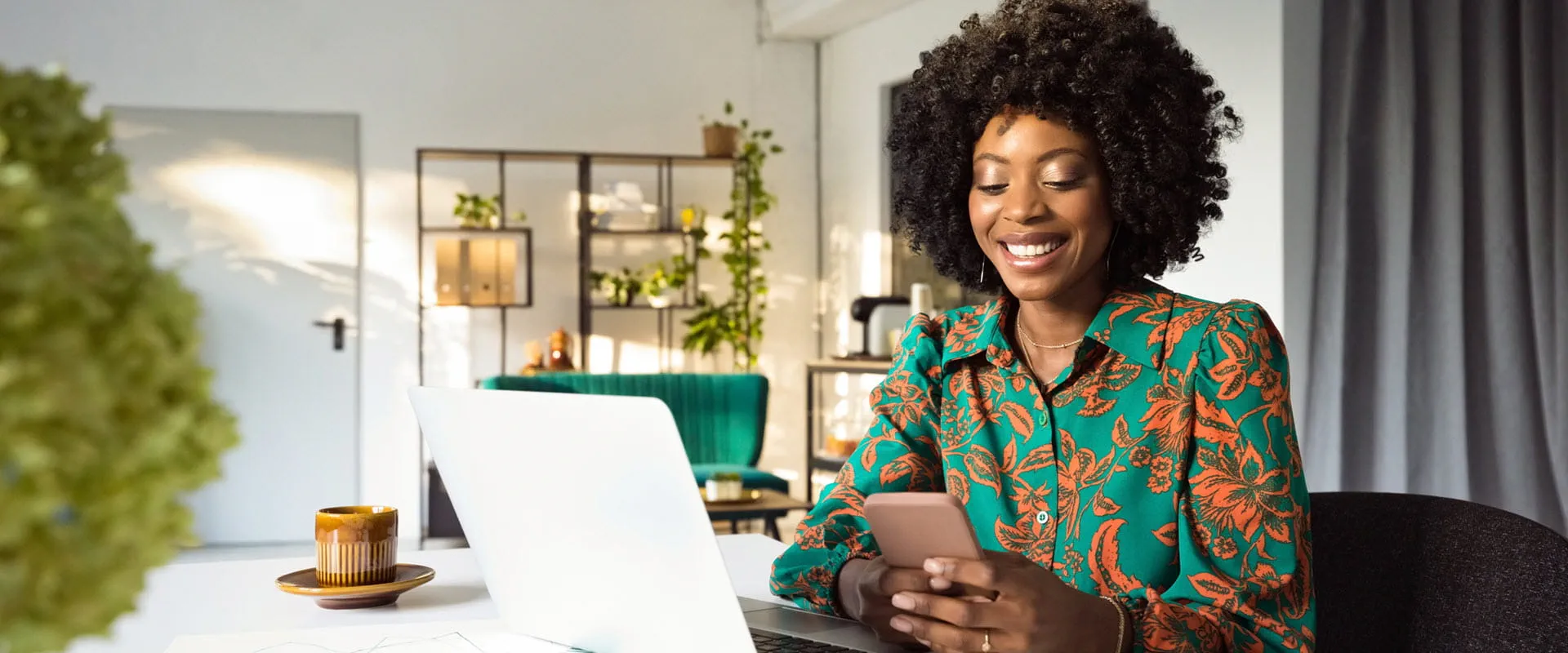 Smiling person in a colorful floral shirt sits at a desk using a smartphone, with a laptop open nearby.