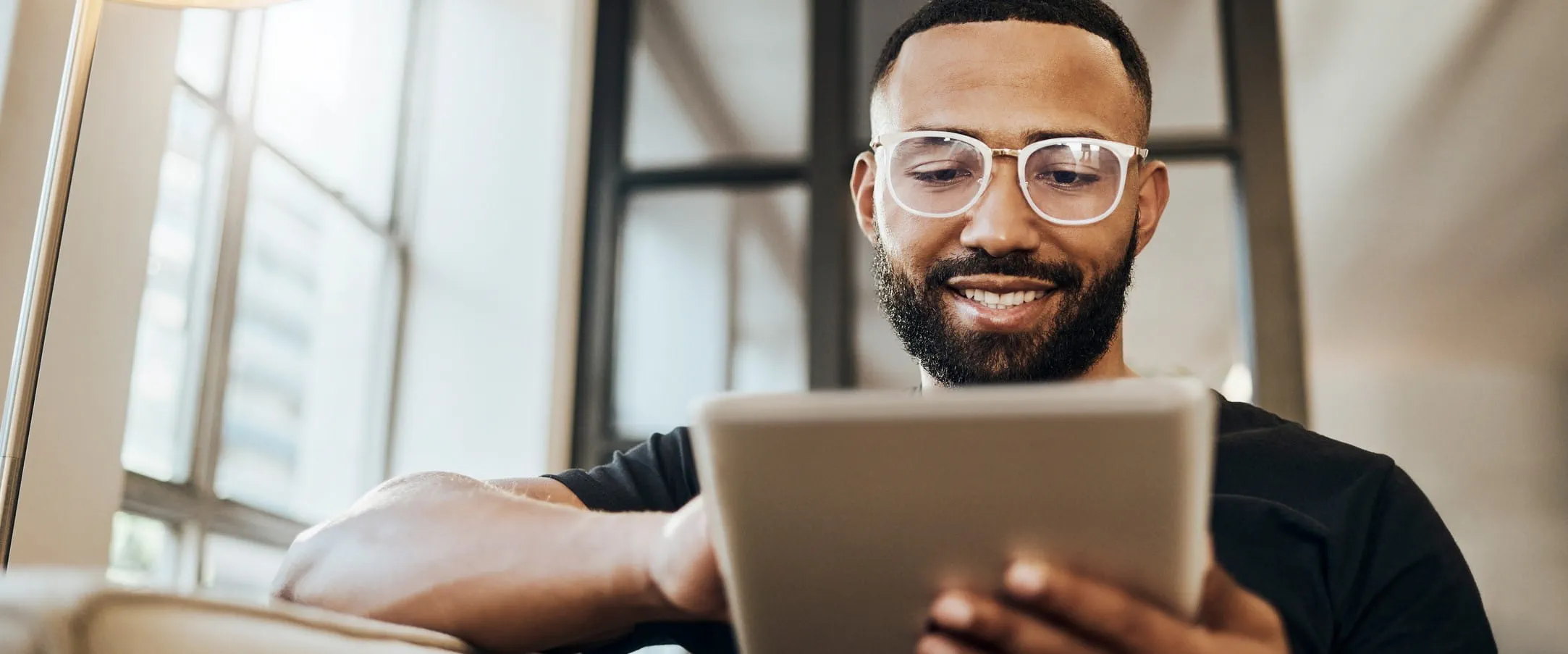 A person wearing glasses smiling while using a tablet in a well-lit room.