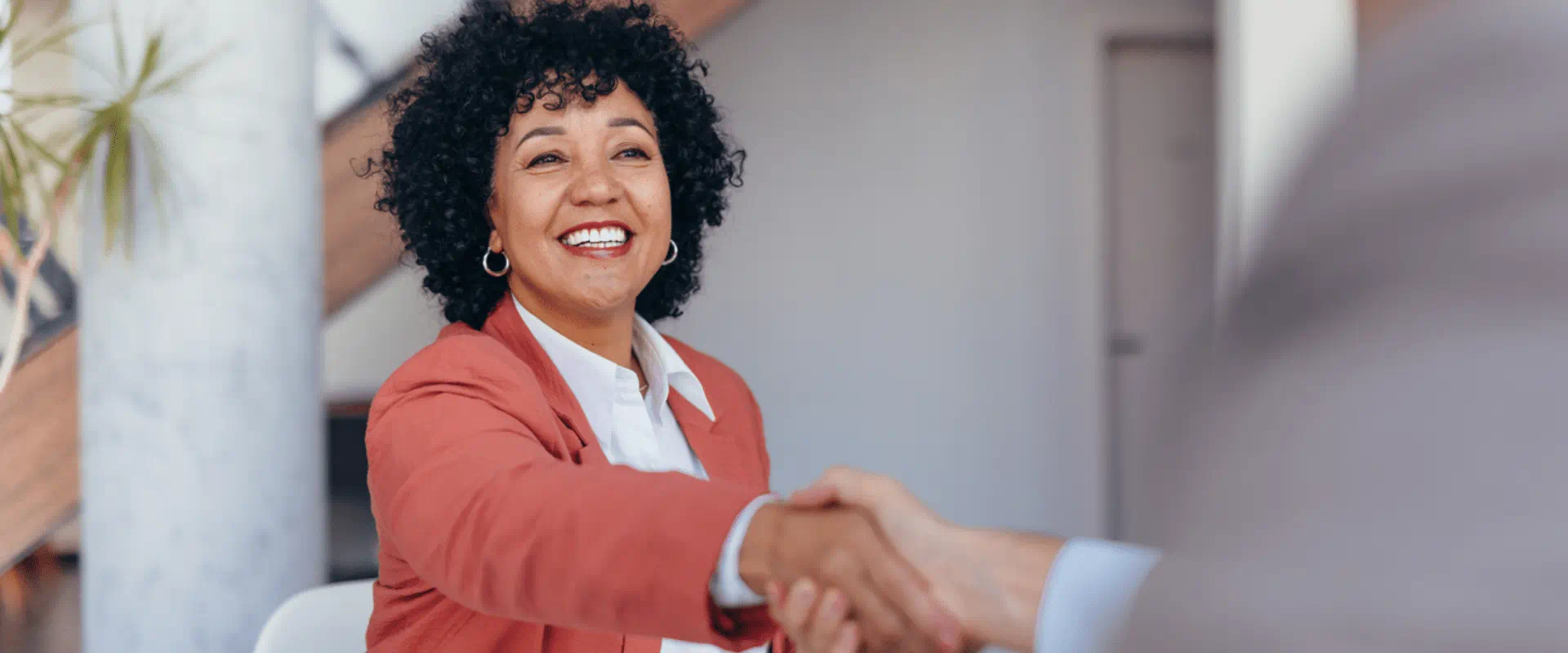 A person interviewing for a job in a big office space, shakes the hand of the person sitting across from her. 