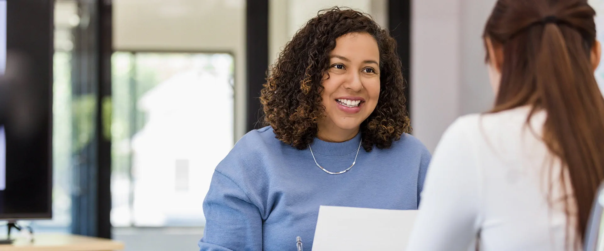 A cheerful woman with curly hair talking to a colleague in a modern office setting, holding a document. A cheerful woman with curly hair talking to a colleague in a modern office setting, holding a document.
